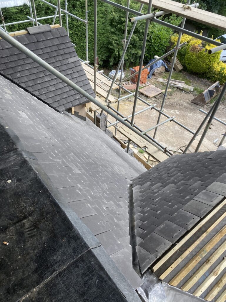 Close view of a slate roof under construction showing valleys, pitched sections, and scaffolding with building materials below.