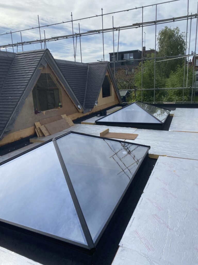 Flat roof under construction with two glass roof lantern skylights installed, surrounded by insulation boards, with scaffolding and dormer roofs in the background.