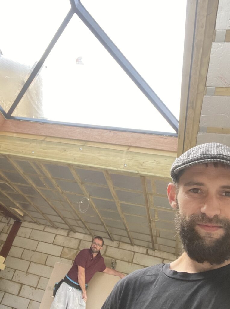 Interior construction scene showing two people, exposed ceiling timbers, insulation boards, and a large triangular glass roof lantern bringing in natural light.