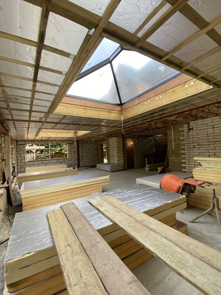 Interior of a building under construction showing exposed wooden ceiling beams, brick walls, stacks of insulation boards, and a large polygonal roof lantern skylight allowing daylight inside.