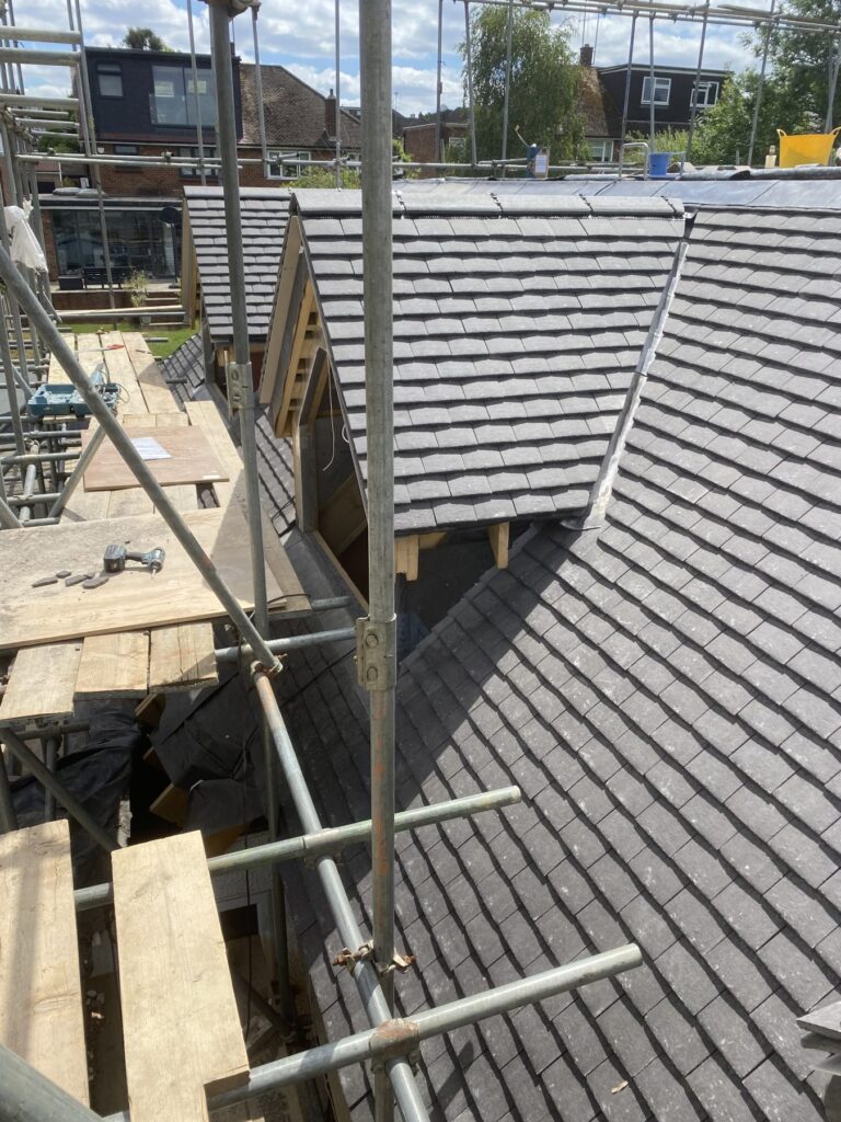 Scaffolding surrounding a pitched roof covered in dark slate tiles during construction, with timber framework and an unfinished dormer window visible.